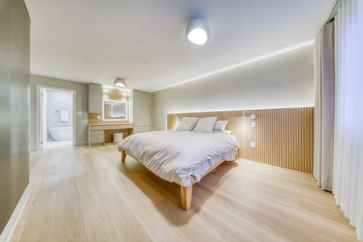 The finished primary bedroom: a slatted oak wall behind the bed and a lit vanity.