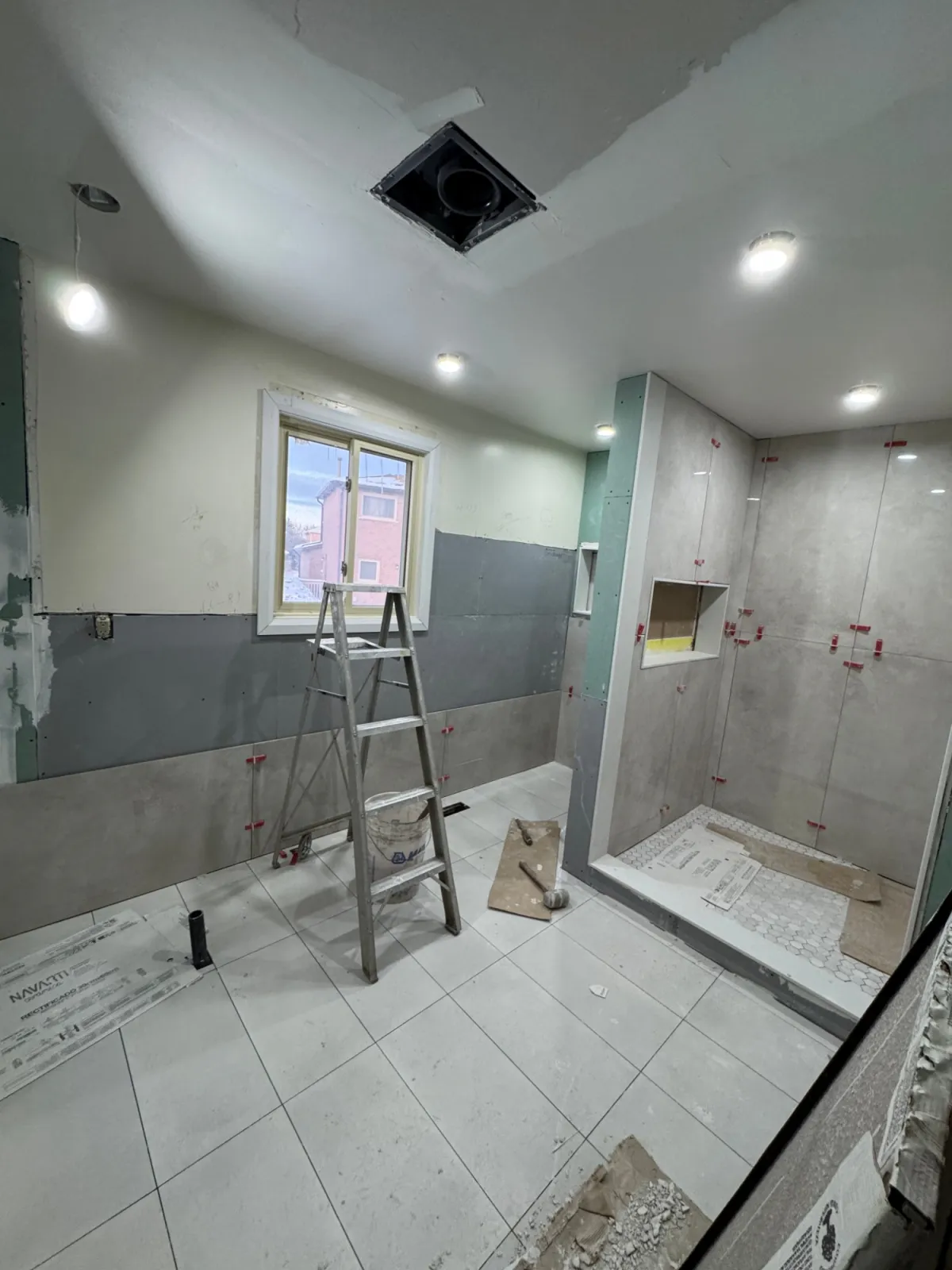 The same ensuite mid-renovation: large grey tile going up with levelling clips, a new shower base and a ladder by the window.
