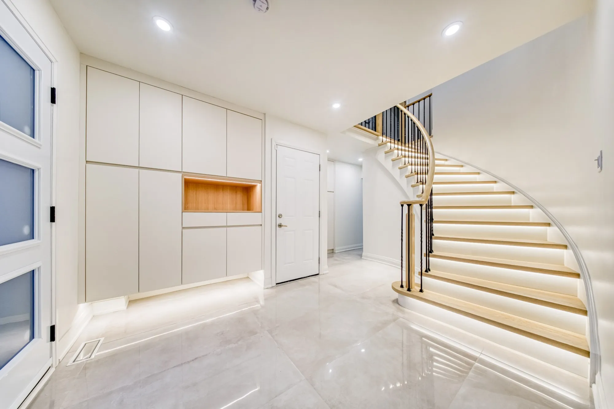 The finished entry: the same curved staircase with oak treads lit from beneath, iron spindles and a wall of built-in storage.